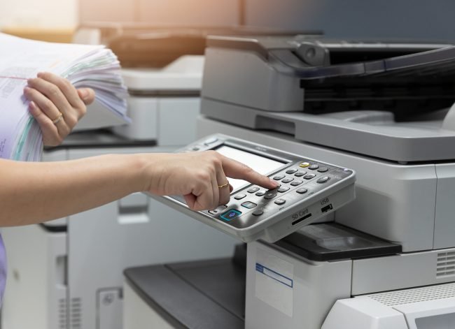 Bussinesswoman using copier machine to copy heap of paperwork in office.